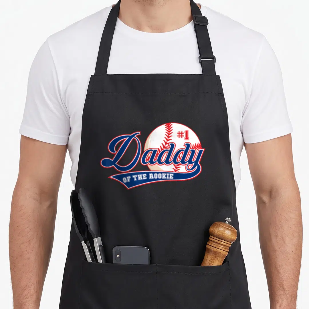 Rookie daddy apron - #1 Daddy OF THE ROOKIE text over a baseball graphic in blue, red, and white — shown on a black cotton canvas apron against a white background