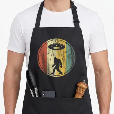 Sasquatch alien bigfoot apron - Retro circular print with vertical stripes, grey walking silhouette, and UFO beaming — shown on a black cotton canvas apron against a white background