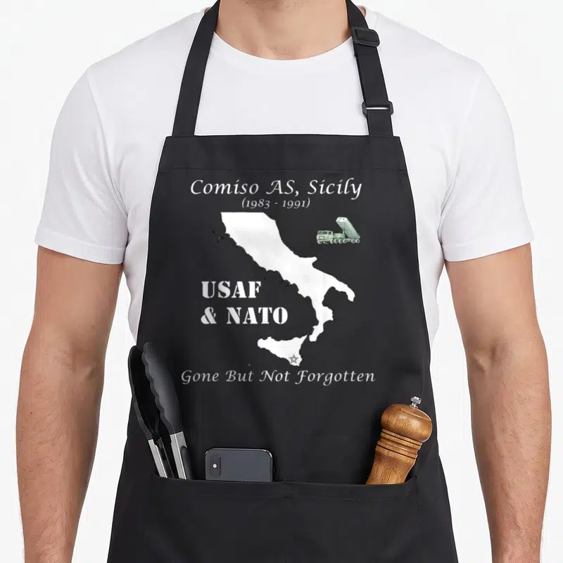Glcm sicily apron - Map of Italy with missile truck and Comiso AS text marking USAF NATO base dates — shown on a black cotton canvas apron against a white background