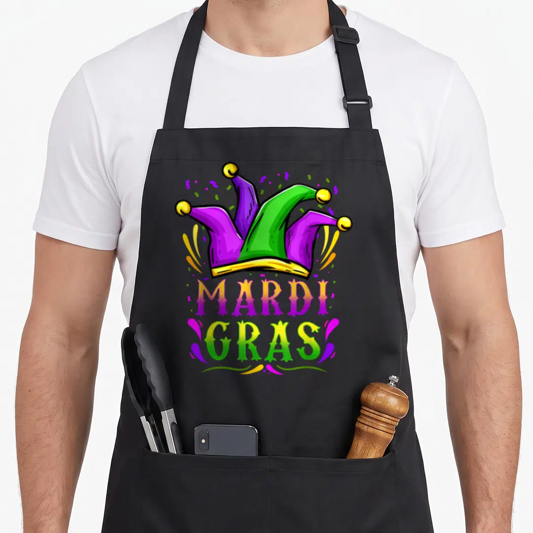 Hat carnival apron - Cartoon jester hat in purple green and yellow with MARDI GRAS text and confetti swirls — shown on a black cotton canvas apron against a white background