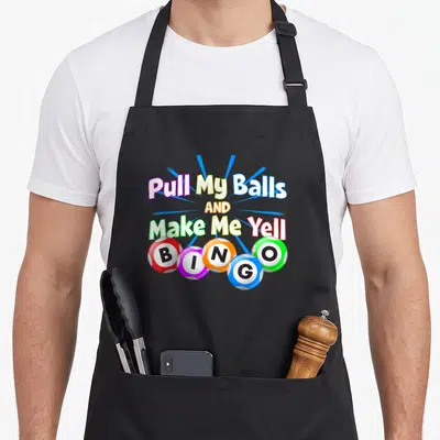 Bingo king apron - Stacked text pull my balls make me yell above five colored bingo balls with blue rays — shown on a black cotton canvas apron against a white background