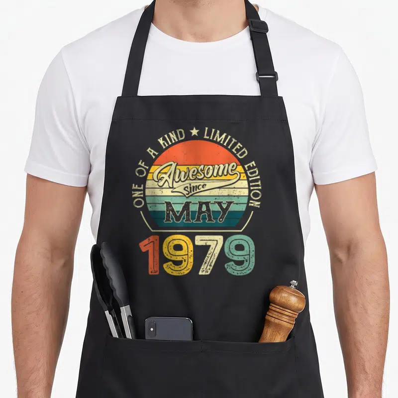 May born day apron - Retro sun graphic with One of a Kind Limited Edition text and colorful 1979 numbers — shown on a black cotton canvas apron against a white background