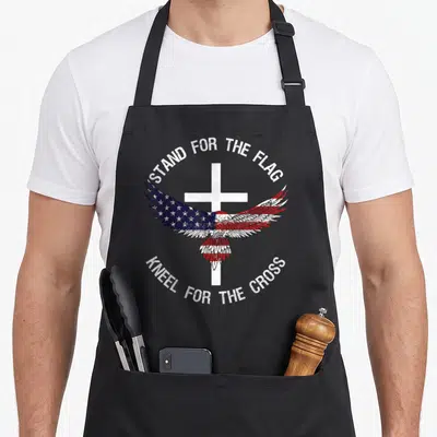 Stand patriotic aprons - Eagle with spread wings over white cross, red white blue distressed text arched above and below — shown on a black cotton canvas apron against a white background