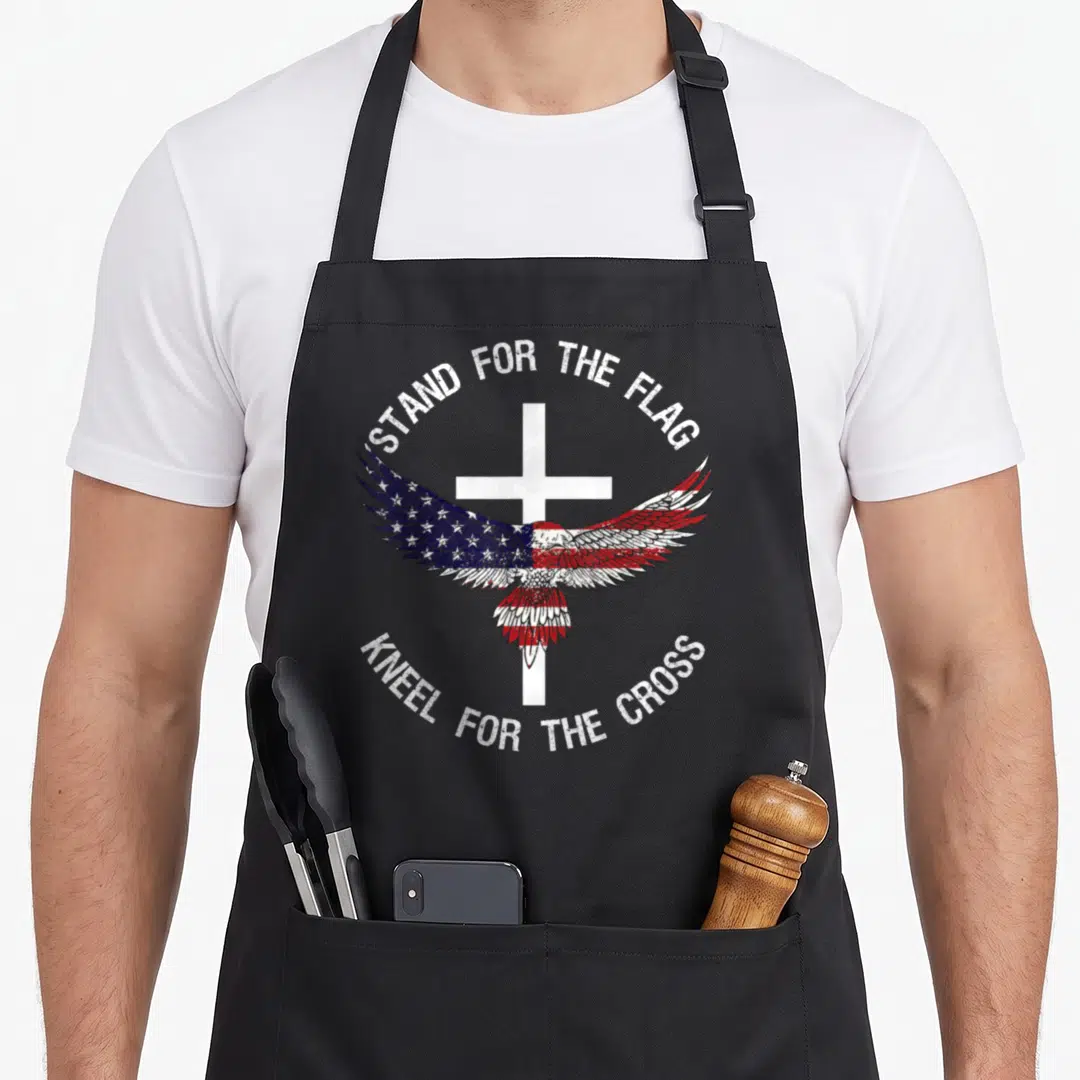 Stand patriotic aprons - Eagle with spread wings over white cross, red white blue distressed text arched above and below — shown on a black cotton canvas apron against a white background