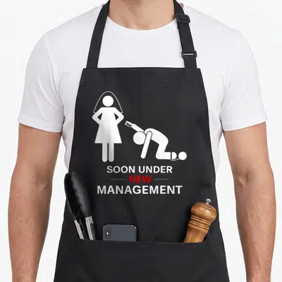 Stag groom apron - White bride silhouette and kneeling groom with ball and chain reading SOON UNDER NEW MANAGEMENT — shown on a black cotton canvas apron against a white background