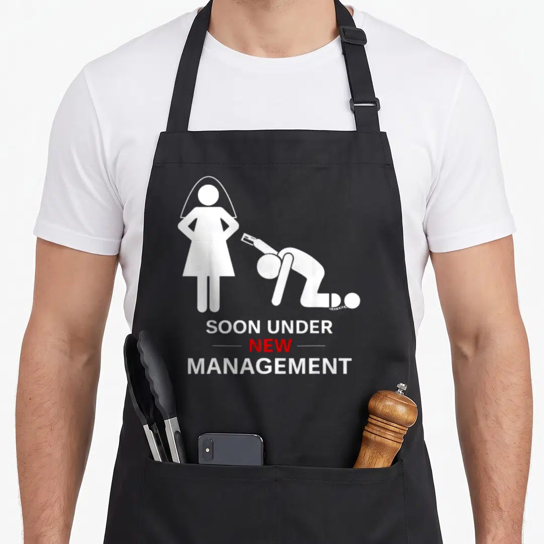 Stag groom apron - White bride silhouette and kneeling groom with ball and chain reading SOON UNDER NEW MANAGEMENT — shown on a black cotton canvas apron against a white background