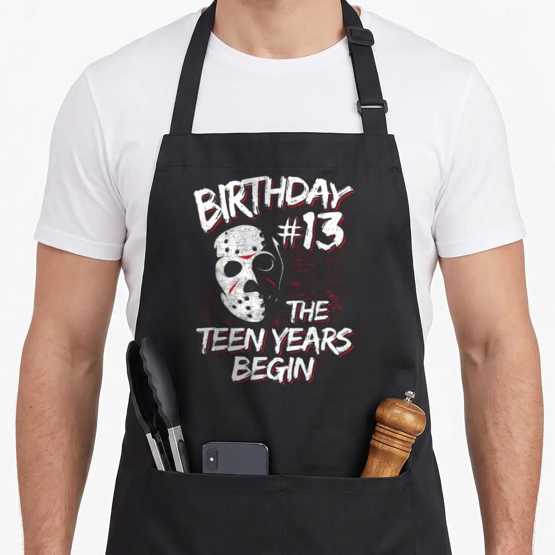 Begin birthday apron - White and red distressed hockey mask with arched text BIRTHDAY #13 THE TEEN YEARS BEGIN — shown on a black cotton canvas apron against a white background