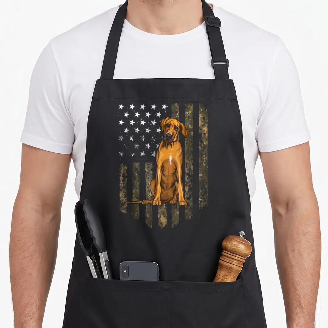 Rhodesian 4th of july apron - Realistic brown dog sitting before tan and green camouflage-patterned USA flag — shown on a black cotton canvas apron against a white background