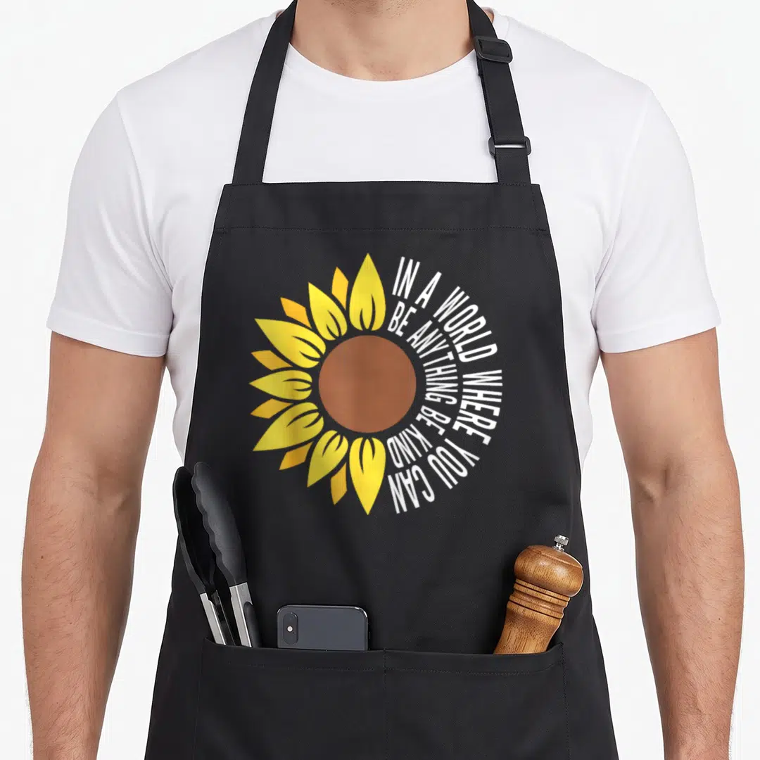 Unity sunflower apron - Yellow sunflower with white text wrapped in circle reading be kind on black fabric — shown on a black cotton canvas apron against a white background