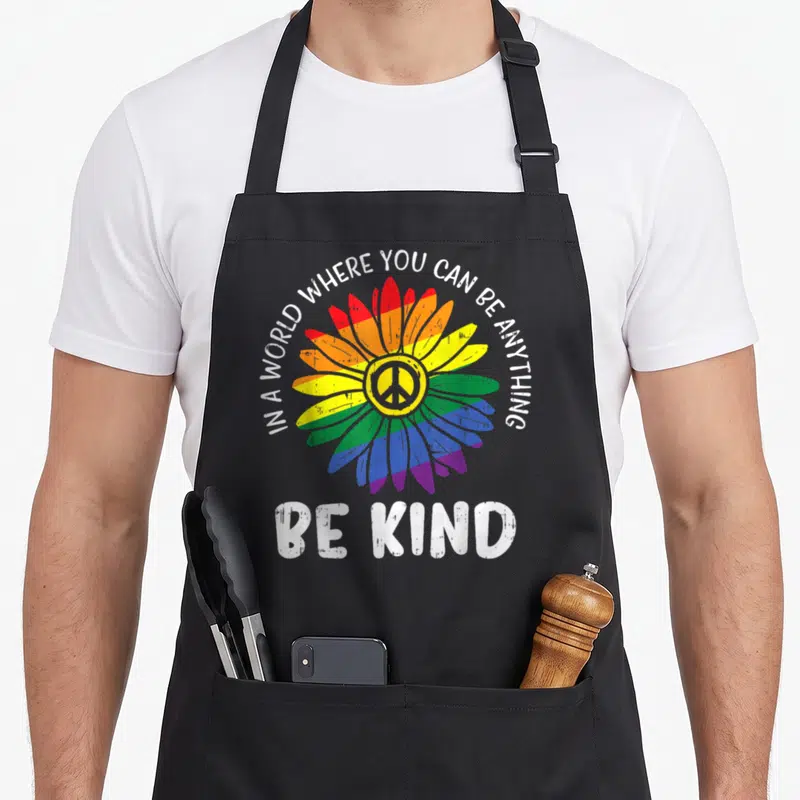 Kind daisy apron - Rainbow daisy flower with peace symbol and white text In a world where you can be anything be kind — shown on a black cotton canvas apron against a white background