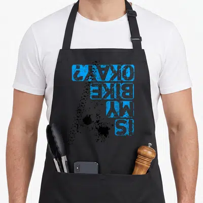 Vintage bike apron - Blue distressed IS MY BIKE OKAY text with black ink splatters and dotted line — shown on a black cotton canvas apron against a white background