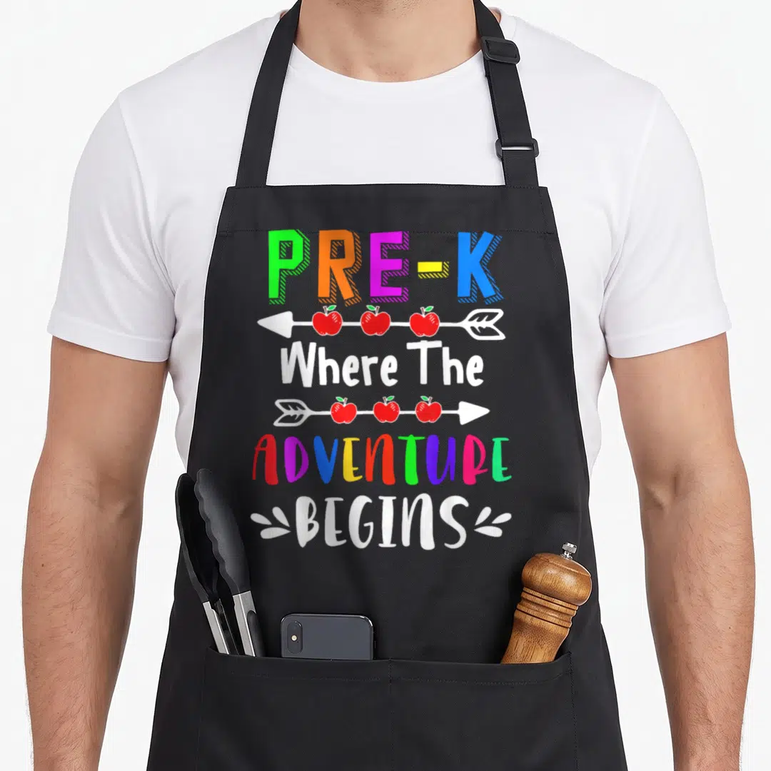 Adventure teacher apron - Stacked PRE-K Where The Adventure Begins text with arrows carrying three red apples each — shown on a black cotton canvas apron against a white background