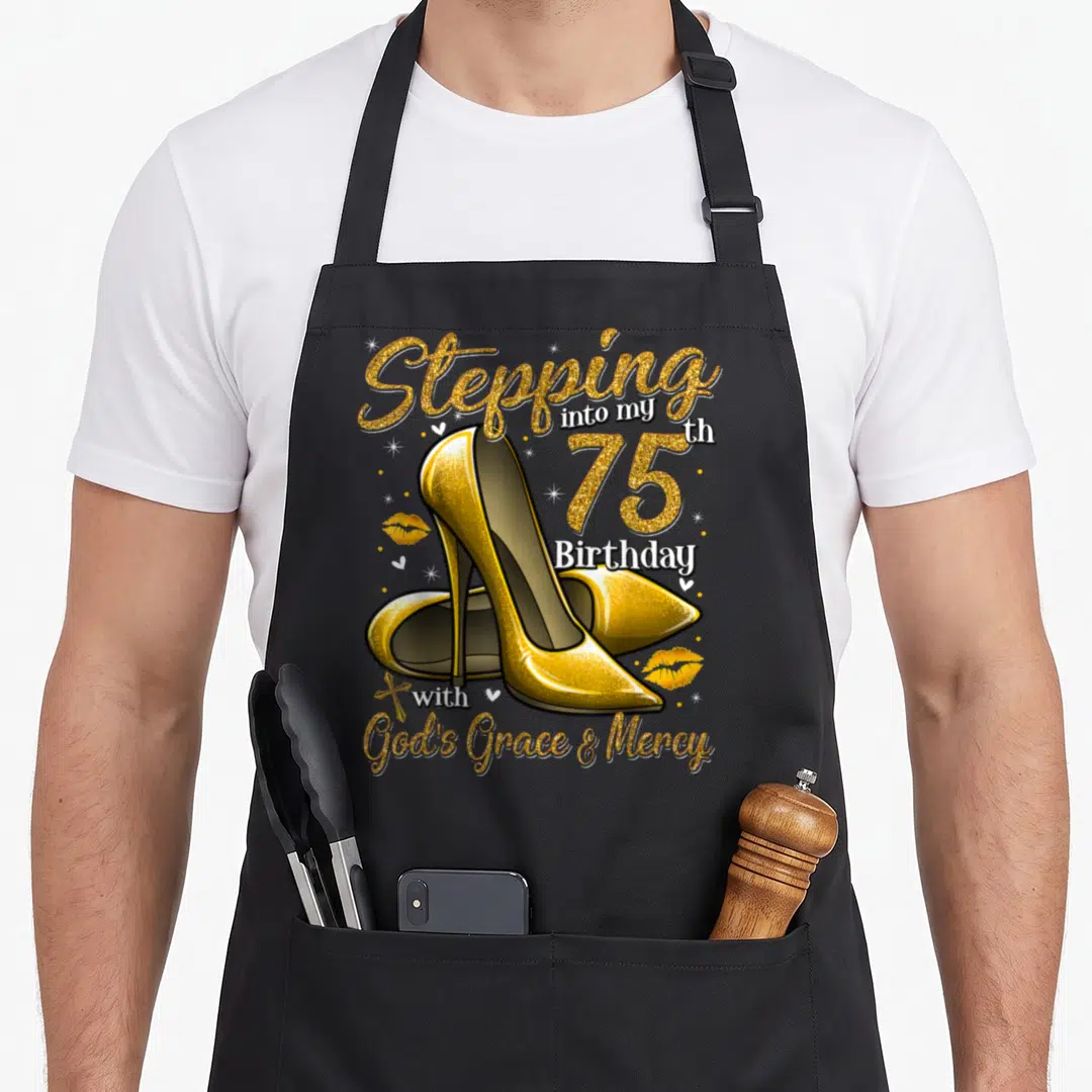 Stepping 75th birthday apron - Gold high heels with text Stepping into my 75th Birthday with God's Grace & Mercy — shown on a black cotton canvas apron against a white background
