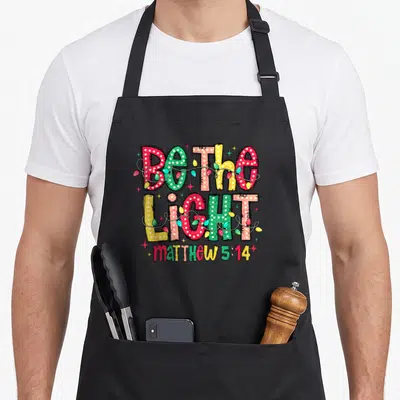 Christian christmas lights apron - Stacked Be The Light text wrapped in red green yellow and blue Christmas lights — shown on a black cotton canvas apron against a white background