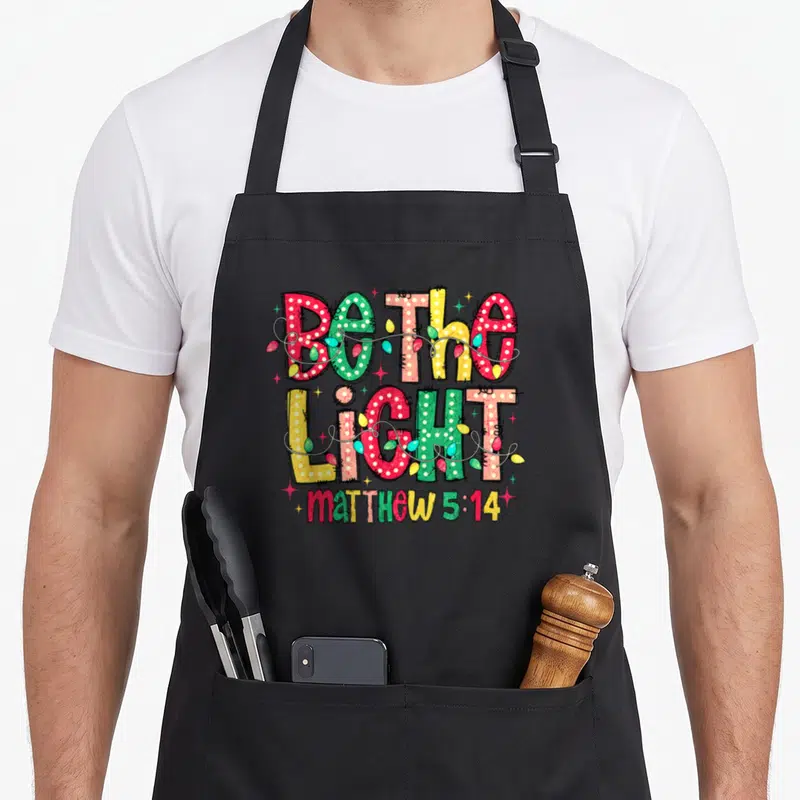 Christian christmas lights apron - Stacked Be The Light text wrapped in red green yellow and blue Christmas lights — shown on a black cotton canvas apron against a white background