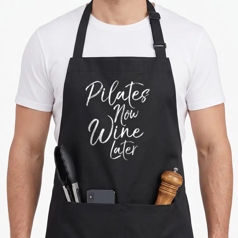 Yoga wine apron - White stacked script text reading Pilates Now Wine Later — shown on a black cotton canvas apron against a white background