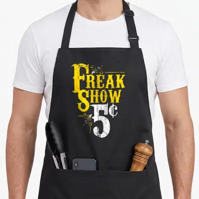 Show weird aprons - Distressed yellow and white text reading Freak Show 5c with decorative swirls — shown on a black cotton canvas apron against a white background