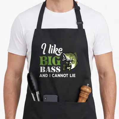 Cannot fishing aprons - Cartoon bass fish next to stacked text 'I like big bass and I cannot lie' in green and white — shown on a black cotton canvas apron against a white background