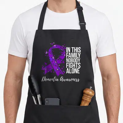 Dementia family aprons - Purple ribbon formed by scattered hearts next to white stacked text and centered script — shown on a black cotton canvas apron against a white background