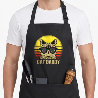 Vintage daddy cat dad apron - Tabby cat wearing black sunglasses in front of a yellow orange and red striped sun — shown on a black cotton canvas apron against a white background