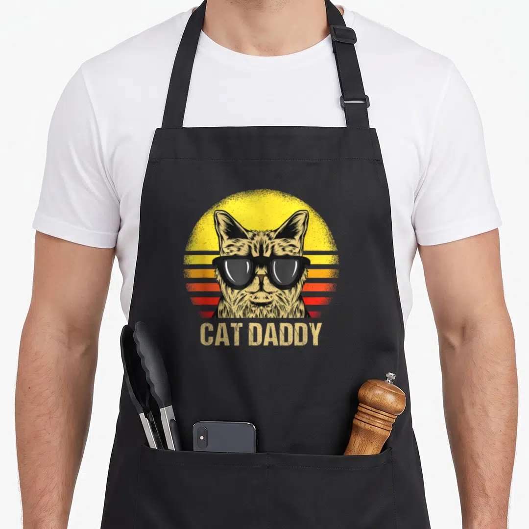 Vintage daddy cat dad apron - Tabby cat wearing black sunglasses in front of a yellow orange and red striped sun — shown on a black cotton canvas apron against a white background