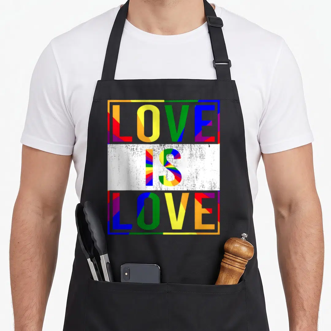 Csd gay pride apron - LOVE IS LOVE text in grey letters filled with red yellow green blue purple geometric rays — shown on a black cotton canvas apron against a white background