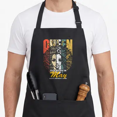May gemini birthday apron - QUEEN WAS BORN IN May HAPPY BIRTHDAY TO ME woman face typography red white blue yellow grey — shown on a black cotton canvas apron against a white background