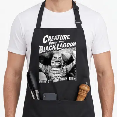 Universal risk apron - Black and white Creature from the Black Lagoon photo with stacked title text and swim warning — shown on a black cotton canvas apron against a white background