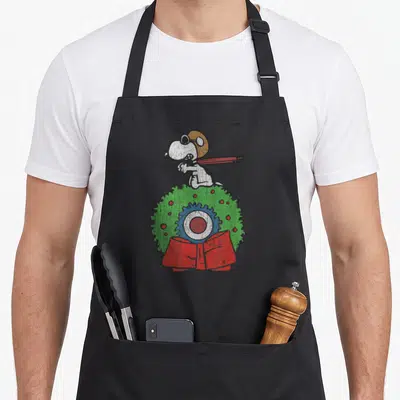 Wreath holiday apron - White cartoon dog standing on green wreath with red bow and berries — shown on a black cotton canvas apron against a white background