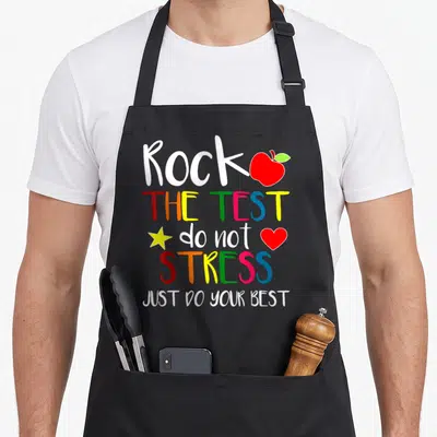 Rock test teacher apron - Stacked colorful text Rock The Test with red apple, yellow star, and heart icons — shown on a black cotton canvas apron against a white background