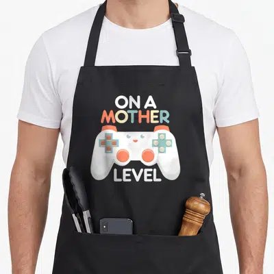Gamer mother's day aprons - White game controller with cute face surrounded by ON A MOTHER LEVEL text in orange and blue — shown on a black cotton canvas apron against a white background
