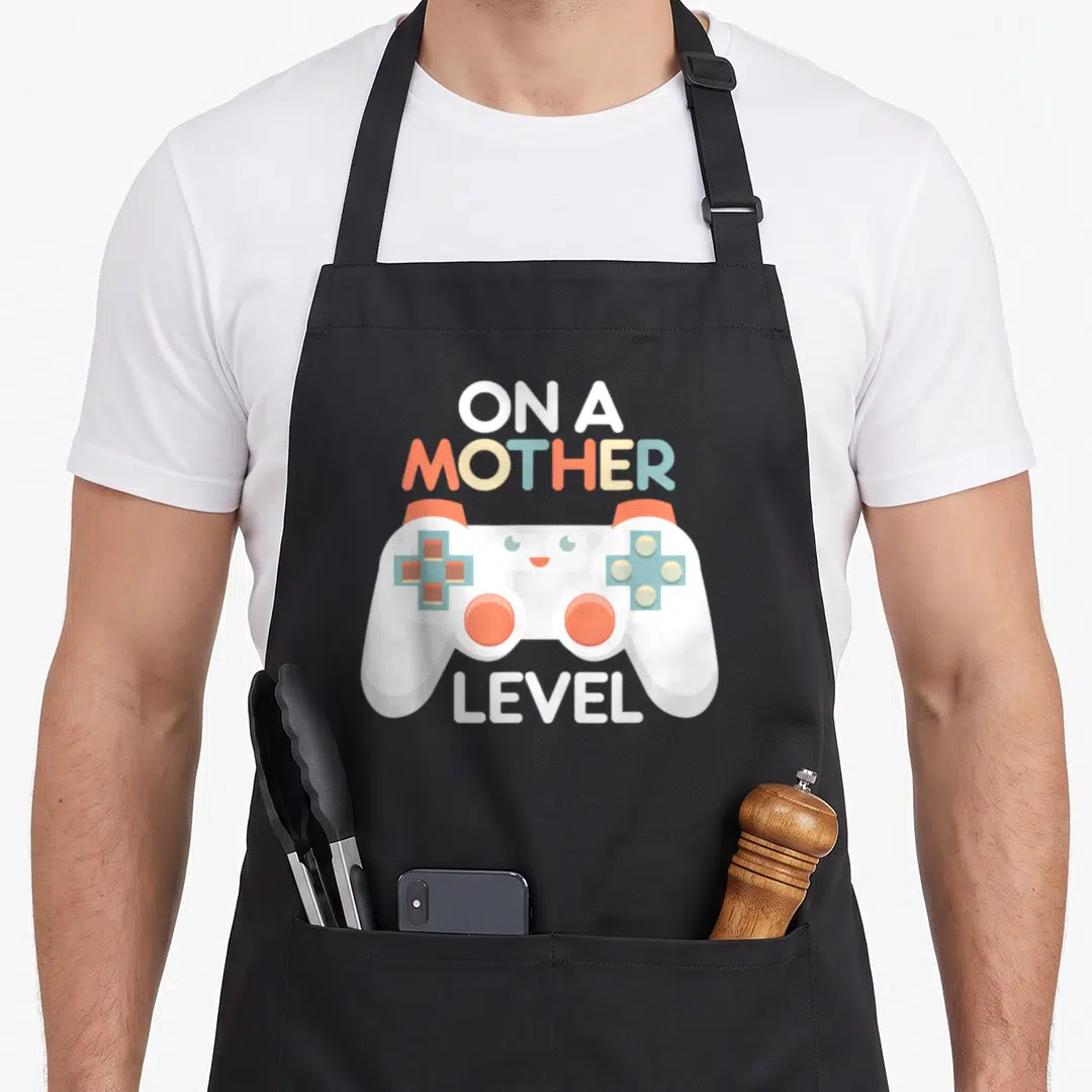 Gamer mother's day aprons - White game controller with cute face surrounded by ON A MOTHER LEVEL text in orange and blue — shown on a black cotton canvas apron against a white background