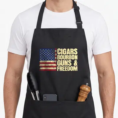 Cigar american flag apron - Distressed US flag beside stacked beige text reading CIGARS BOURBON GUNS & FREEDOM — shown on a black cotton canvas apron against a white background