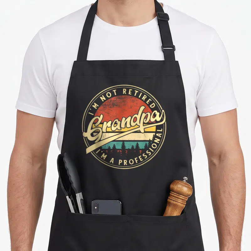 Grandparent professional aprons - Retro circular badge with Grandpa text over sunset and forest silhouette in beige red — shown on a black cotton canvas apron against a white background