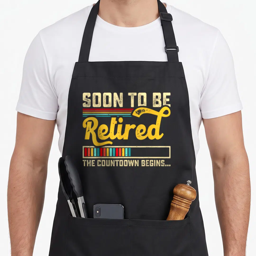 Begin retirement apron - Retro text Soon To Be Retired with striped progress bar in beige yellow red teal blue — shown on a black cotton canvas apron against a white background