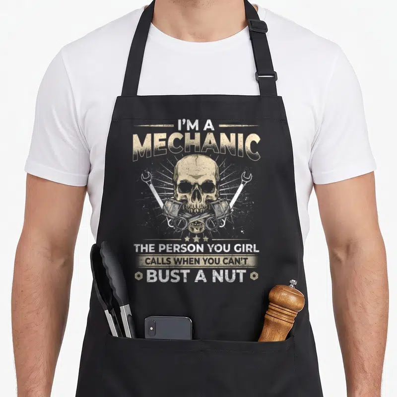 Vintage car auto mechanic apron - Tan distressed skull between crossed wrenches and pistons with five stars below — shown on a black cotton canvas apron against a white background