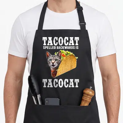 Tacocat cat apron - Cat head inside a taco with TACOCAT text above and below on grey background — shown on a black cotton canvas apron against a white background