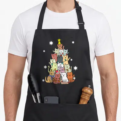 Ugly xmas apron - Stacked cartoon cats forming a Christmas tree with string lights and a star on top — shown on a black cotton canvas apron against a white background