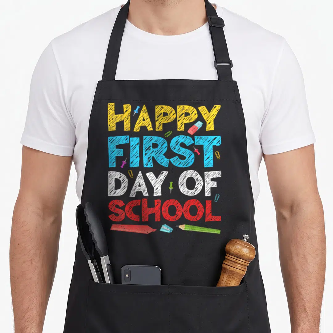School day family apron - Stacked Happy First Day of School text in yellow blue white red with eraser pencil — shown on a black cotton canvas apron against a white background