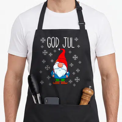 God scandinavian aprons - Cartoon gnome in red hat and blue coat below white GOD JUL text and snowflakes — shown on a black cotton canvas apron against a white background