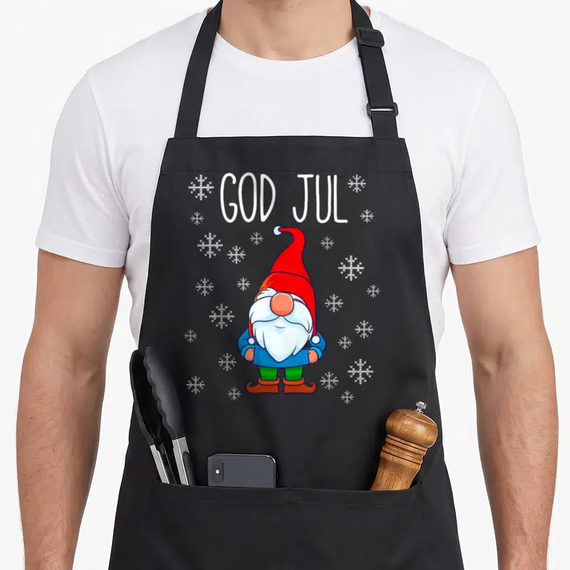 God scandinavian aprons - Cartoon gnome in red hat and blue coat below white GOD JUL text and snowflakes — shown on a black cotton canvas apron against a white background