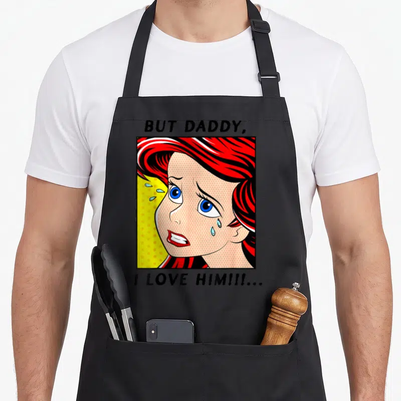 Ariel daddy apron - Comic style woman with red hair crying in a yellow dotted square with text above and below — shown on a black cotton canvas apron against a white background