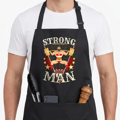 Squat strongman apron - Vintage strongman lifting barbell with STRONG MAN text and sunburst stars in beige red gold — shown on a black cotton canvas apron against a white background