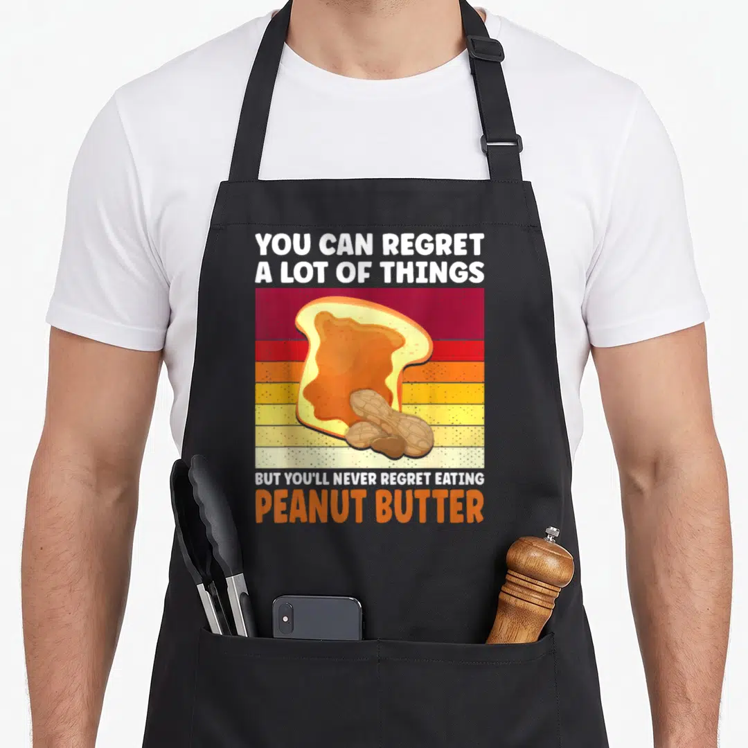 Youll butter apron - Retro striped toast with peanut butter and two peanuts in shells under white and orange text — shown on a black cotton canvas apron against a white background