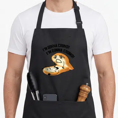 Crumb punny apron - Arched 'I'M GONNA CRUMB!' text above cartoon bread split open with angry and laughing faces — shown on a black cotton canvas apron against a white background