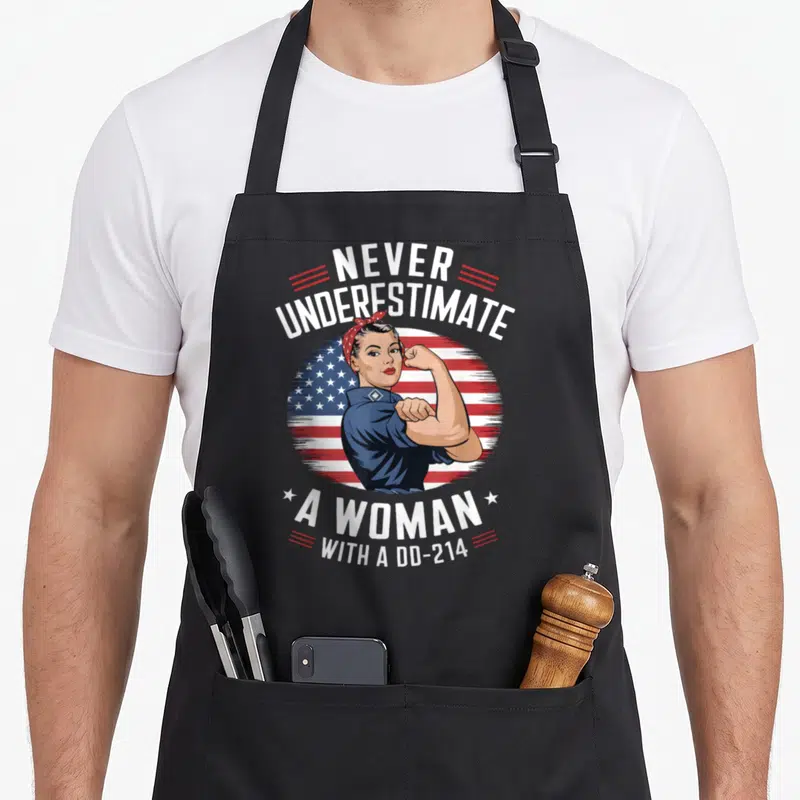 Funny underestimate female veteran apron - Retro woman flexing arm in front of circular US flag with white and red text — shown on a black cotton canvas apron against a white background