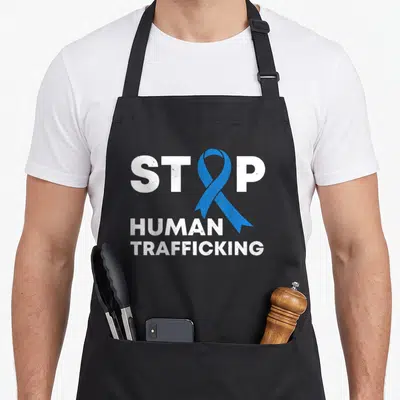 Trafficking ribbon apron - White distressed text STOP HUMAN TRAFFICKING with blue awareness ribbon replacing the letter — shown on a black cotton canvas apron against a white background