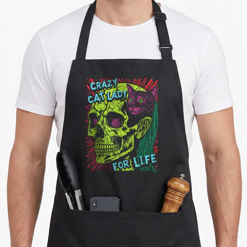 Goth crazy cat lady apron - Lime skull with pink cat overlap and blue text surrounded by red lines — shown on a black cotton canvas apron against a white background