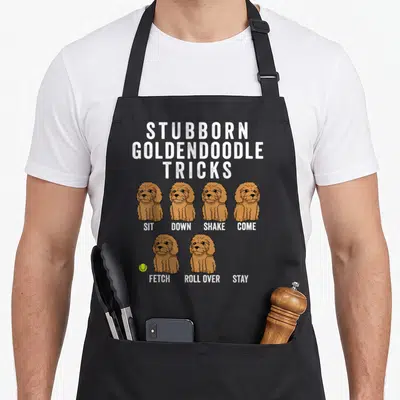 Stubborn goldendoodle apron - Six brown cartoon dogs with trick labels and a yellow-green tennis ball — shown on a black cotton canvas apron against a white background