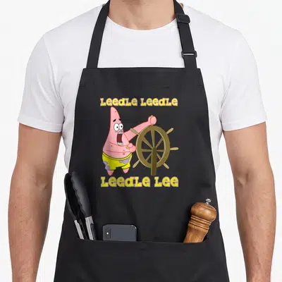 Leedle patrick apron - Patrick Star holding brown ship wheel with yellow LeedLe text — shown on a black cotton canvas apron against a white background